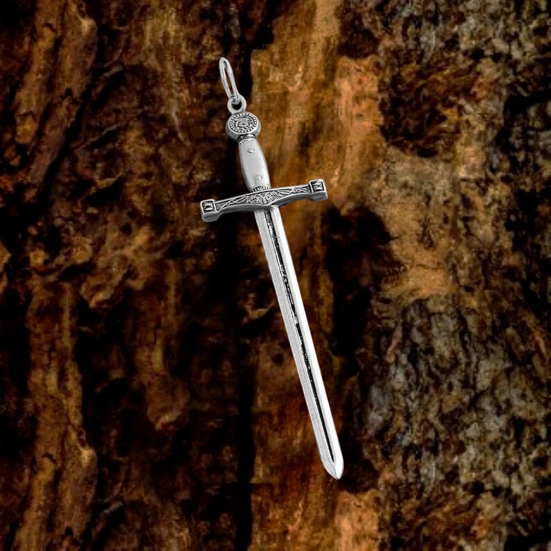 May include: Silver sword pendant with intricate details. The sword has a pointed tip and a cross guard. The pendant is hanging from a chain.