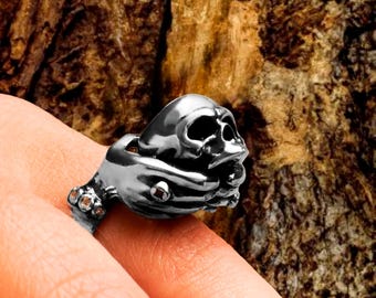 Puede incluir: Un anillo de plata con un par de manos sosteniendo una calavera. El anillo está detallado con superficies texturizadas y pequeños tachuelas.