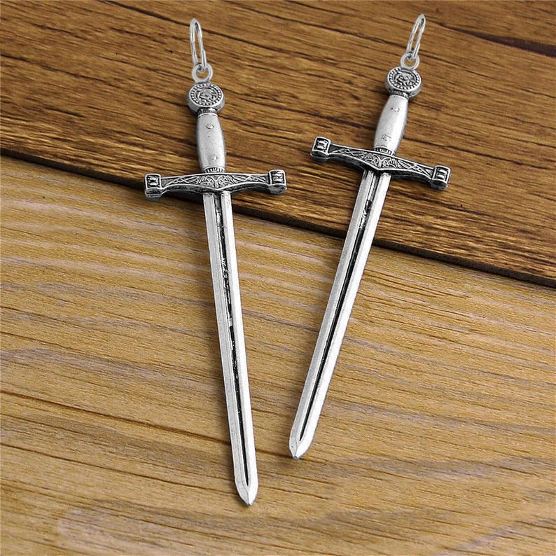 May include: Two silver sword pendant charms with intricate details. The swords have a pointed tip and a cross guard.