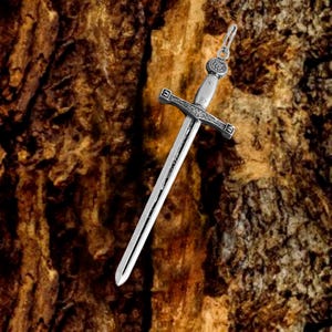 May include: A silver sword pendant with intricate details and a cross-shaped hilt. The pendant is hanging against a brown wooden background.