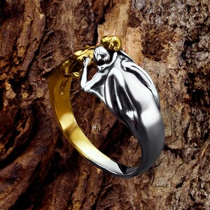 May include: A silver ring with a gold accent features a sculpted design of two figures embracing. The ring is displayed on a textured wooden surface.