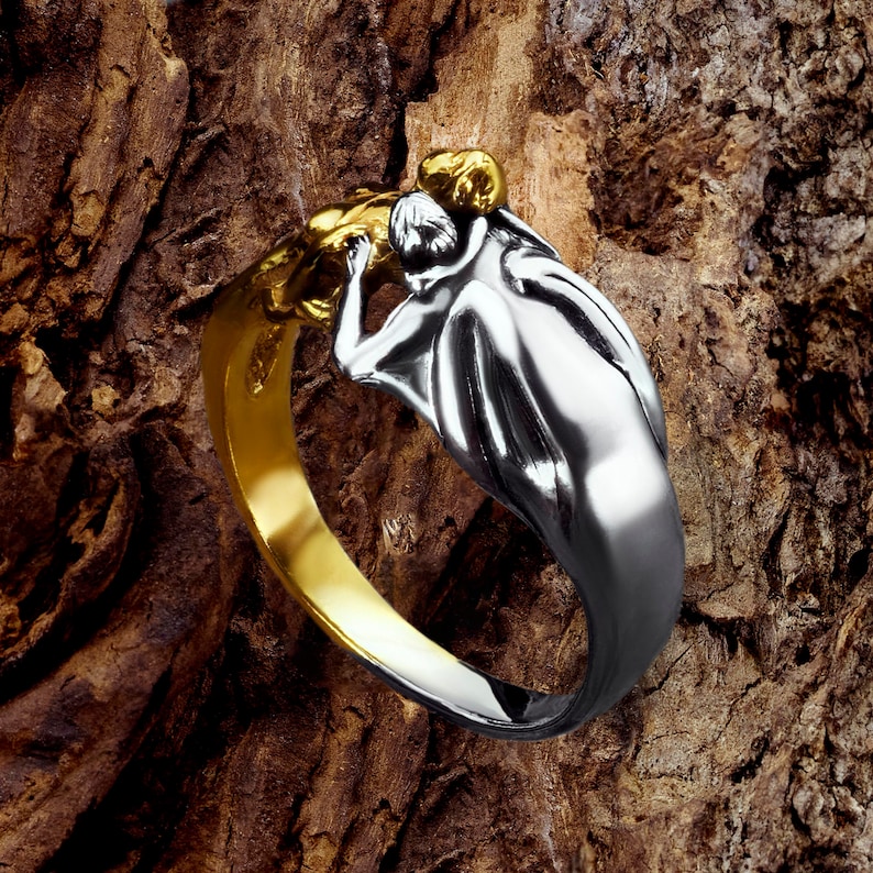 May include: A silver ring with a gold accent features a sculpted design of two figures embracing. The ring is displayed on a textured wooden surface.