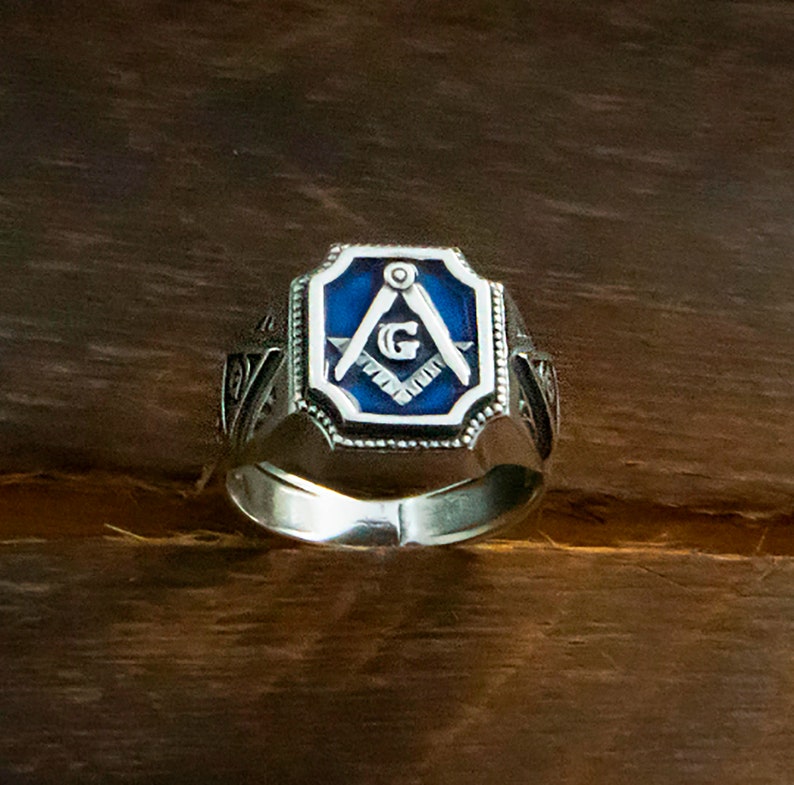 Op de afbeelding: Een zilveren ring met een blauw ge&euml;mailleerd vierkant met een zilveren kompas en de letter G. De ring heeft ingewikkelde details op de band.