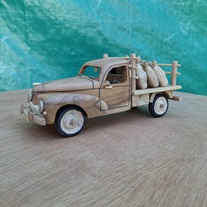 Può includere: Un modellino di camion in legno realizzato a mano con una finitura in venature di legno naturale color marrone chiaro. Il veicolo presenta un pianale di carico con tre piccoli sacchi di legno arrotondati, quattro ruote bianche e una cabina.