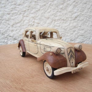 May include: A wooden model of a classic car, featuring a light beige body and brown fenders. The car has detailed features like headlights, wheels, and a front bumper with the text "2025 PAM". It is displayed on a wooden surface.