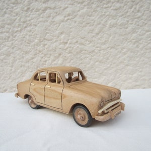 Può includere: Un'auto modello in legno di colore chiaro con quattro porte, un cofano e un paraurti anteriore. L'auto ha quattro ruote e sembra essere un modello d'epoca. L'auto è in legno e ha una finitura naturale.