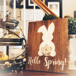 May include: A wooden sign with a white bunny silhouette and the text "Hello Spring!" The bunny has a burlap bow with a white lace flower on its tail.