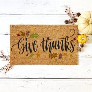 May include: A brown coir doormat with the text "Give Thanks" in black cursive lettering. The doormat is decorated with fall-colored leaves.