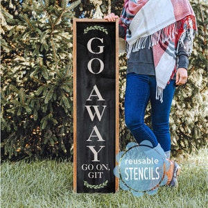 May include: A black wooden sign with white lettering that reads "GO AWAY GO ON, GIT". The sign is decorated with green leaves at the top and bottom. The sign is being held by a person wearing blue jeans and a red and white plaid scarf. The sign is in front of a green grassy area.