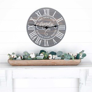 Puede incluir: Un reloj de madera gris con n&uacute;meros romanos y el texto "El tiempo pasado con la familia vale cada minuto". El reloj est&aacute; colgado en una pared blanca sobre una bandeja de madera llena de tallos de algod&oacute;n y eucalipto.