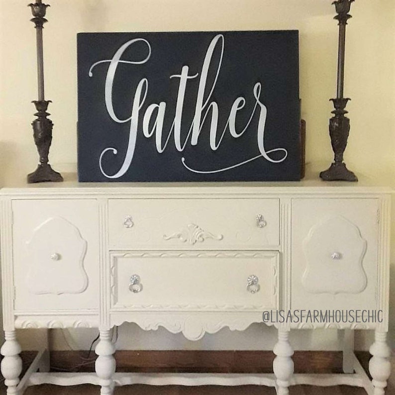 May include: A white painted antique buffet with ornate carvings and silver knobs. A black sign with the word "Gather" in silver script is displayed on top of the buffet.