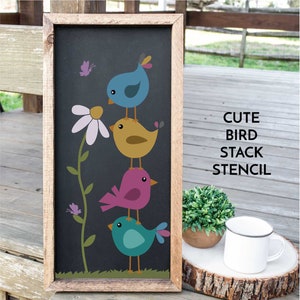 May include: A wooden sign with a black background featuring a stack of four colorful birds. The birds are perched on top of each other, with a yellow bird at the top, a pink bird in the middle, a blue bird at the bottom, and a teal bird in the middle. The sign also features a white daisy with a green stem and two small butterflies. The text "CUTE BIRD STACK STENCIL" is written in white letters on the right side of the sign.