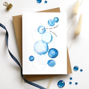 May include: Greeting card with watercolor blue bubbles and the text "May all your troubles be bubbles." The card is on a brown envelope, with a navy blue ribbon, gold clip, and blue gems scattered around.
