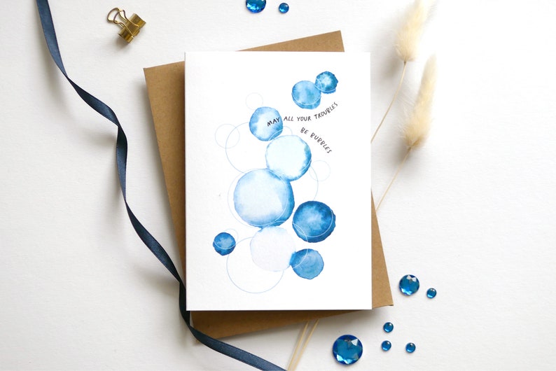 May include: Greeting card with watercolor blue bubbles and the text "May all your troubles be bubbles." The card is on a brown envelope, with a navy blue ribbon, gold clip, and blue gems scattered around.