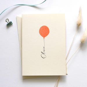 May include: A blank card with a simple design of a single orange balloon with a string that says "Olivia" in black cursive.