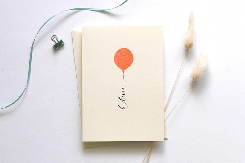 May include: A blank card with a simple design of a single orange balloon with a string that says "Olivia" in black cursive.