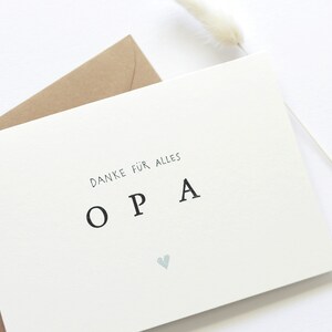Op de afbeelding: Een witte kaart met de tekst "Danke f&uuml;r alles OPA" in zwarte letters en een klein blauw hartje. De kaart ligt op een bruine envelop.