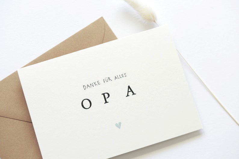 Op de afbeelding: Een witte kaart met de tekst "Danke f&uuml;r alles OPA" in zwarte letters en een klein blauw hartje. De kaart ligt op een bruine envelop.
