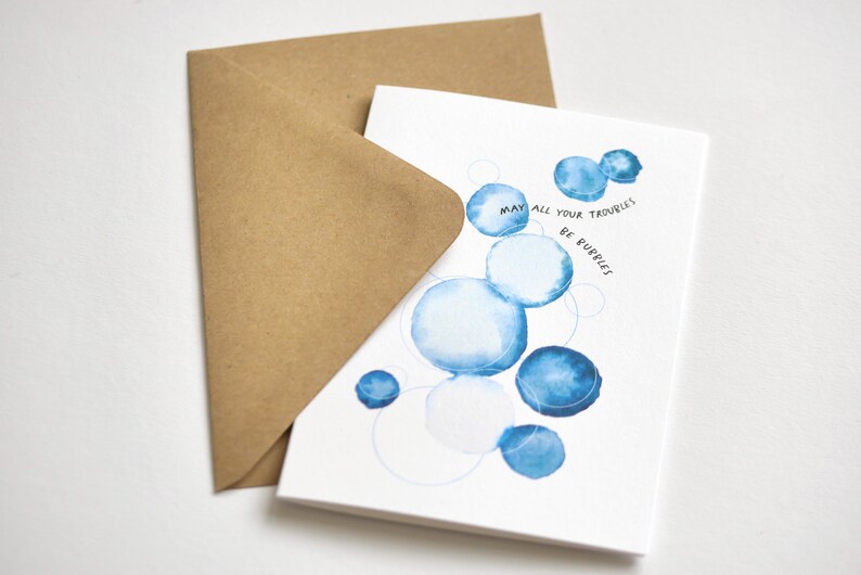 May include: A greeting card with blue watercolor bubbles and the text "May all your troubles be bubbles." The card is paired with a brown envelope. The design is simple and elegant, suitable for various occasions.