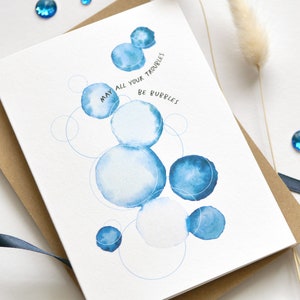 May include: Greeting card with watercolor blue bubble design and the text "MAY ALL YOUR TROUBLES BE BUBBLES". The card is on a brown envelope with a blue ribbon and blue gems.