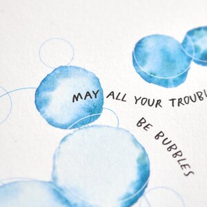 May include: Watercolor art print featuring blue bubble shapes and the handwritten text "MAY ALL YOUR TROUBLES BE BUBBLES." The artwork has a soft, airy feel with delicate lines and watercolor washes, perfect for home decor.