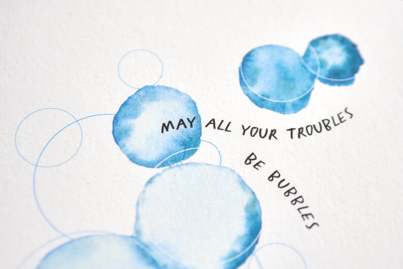 May include: Watercolor art print featuring blue bubble shapes and the handwritten text "MAY ALL YOUR TROUBLES BE BUBBLES." The artwork has a soft, airy feel with delicate lines and watercolor washes, perfect for home decor.