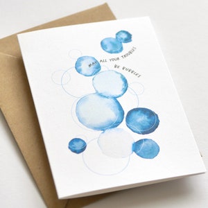 May include: Greeting card with a watercolor design of blue bubbles and the text "May all your troubles be bubbles." The card is paired with a brown envelope. The design features various sizes of blue circles.
