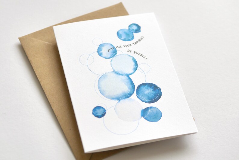 May include: Greeting card with a watercolor design of blue bubbles and the text "May all your troubles be bubbles." The card is paired with a brown envelope. The design features various sizes of blue circles.