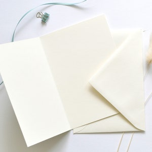 May include: A blank white card with a folded edge, a white envelope, and two dried white pampas grass stems on a white background.