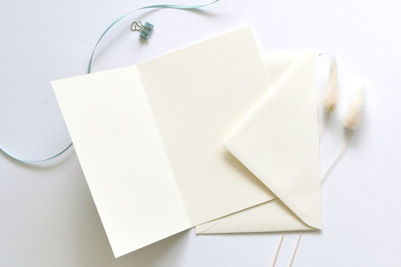 May include: A blank white card with a folded edge, a white envelope, and two dried white pampas grass stems on a white background.