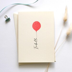May include: A blank card with a red balloon and the word "Isabella" written in black calligraphy. The card is on a white background with a blue ribbon and dried pampas grass.