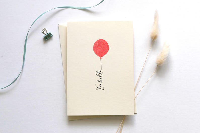 May include: A blank card with a red balloon and the word "Isabella" written in black calligraphy. The card is on a white background with a blue ribbon and dried pampas grass.