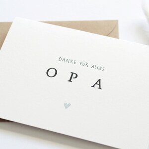 Op de afbeelding: Een witte kaart met de tekst "Danke f&uuml;r alles OPA" en een klein blauw hartje. De kaart ligt op een bruine envelop.