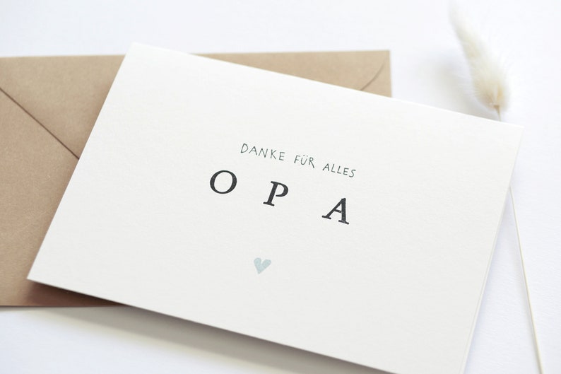 Op de afbeelding: Een witte kaart met de tekst "Danke f&uuml;r alles OPA" en een klein blauw hartje. De kaart ligt op een bruine envelop.