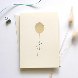 May include: A blank card with a gold glitter balloon and the word "Happy" written in black calligraphy.