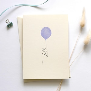 May include: A blank card with a purple balloon and the word "Lila" written in cursive. The card is on a white surface with a blue ribbon and dried pampas grass.