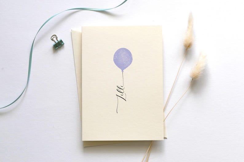 May include: A blank card with a purple balloon and the word "Lila" written in cursive. The card is on a white surface with a blue ribbon and dried pampas grass.