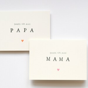 May include: Two white cards with the words "Danke fur alles" and "PAPA" on one card and "MAMA" on the other card. Each card has a small heart in a different color.