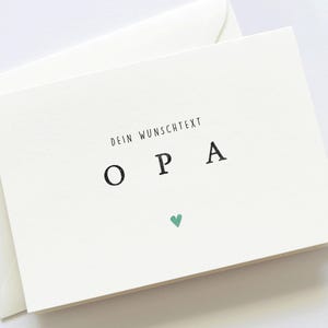 Op de afbeelding: Een witte kaart met zwarte tekst die "Dein Wunschtext OPA" zegt met een klein groen hart onder de tekst.