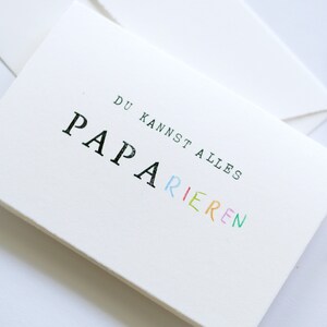 以下が含まれることがあります： 白地に黒とカラフルな文字で「DU KANNST ALLES PAPA RIEREN」と書かれたカード。 「PAPA」という単語は虹色です。
