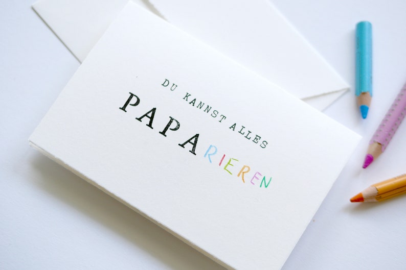以下が含まれることがあります： 白地に黒とカラフルな文字で「DU KANNST ALLES PAPA RIEREN」と書かれたカード。 「PAPA」という単語は虹色です。