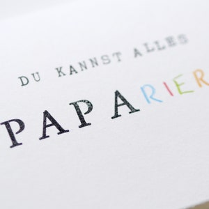 以下が含まれることがあります： 白いカードに「DU KANNST ALLES PAPARIEREN」という黒い文字とカラフルな文字が書かれています。 「PAPARIEREN」という単語は虹色で書かれています。