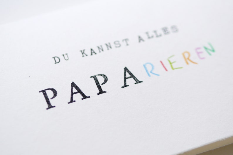 以下が含まれることがあります： 白いカードに「DU KANNST ALLES PAPARIEREN」という黒い文字とカラフルな文字が書かれています。 「PAPARIEREN」という単語は虹色で書かれています。