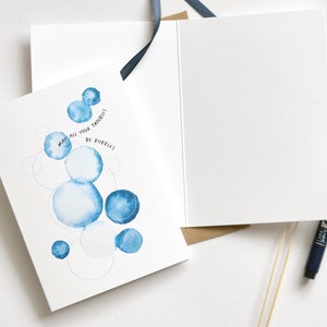 May include: Greeting card with a watercolor design of blue bubbles and the text "May all your troubles be bubbles." The card is open, revealing a blank interior. A black pen and blue ribbon are also visible.