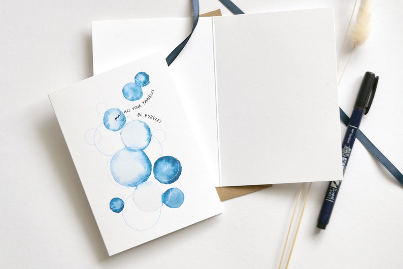 May include: Greeting card with a watercolor design of blue bubbles and the text "May all your troubles be bubbles." The card is open, revealing a blank interior. A black pen and blue ribbon are also visible.