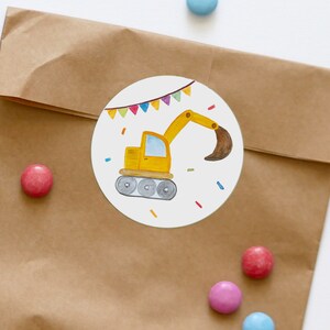 May include: A round sticker featuring a yellow construction vehicle on a white background. The vehicle has a brown scoop and is surrounded by colourful confetti and a string of flags.