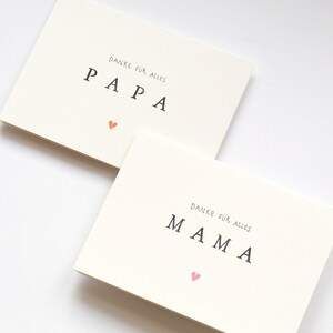May include: Two white cards with the words "PAPA" and "MAMA" written in black. The word "PAPA" has a small orange heart below it and the word "MAMA" has a small pink heart below it. The text above each word reads "Danke fur alles".