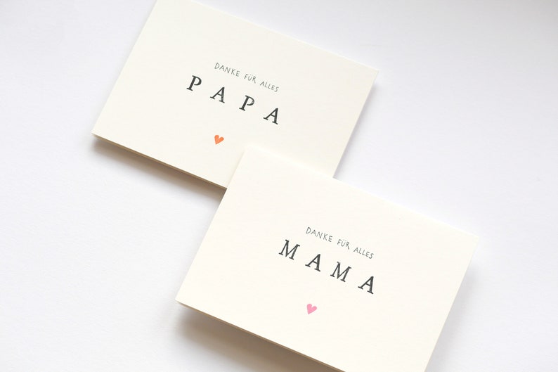 May include: Two white cards with the words "PAPA" and "MAMA" written in black. The word "PAPA" has a small orange heart below it and the word "MAMA" has a small pink heart below it. The text above each word reads "Danke fur alles".