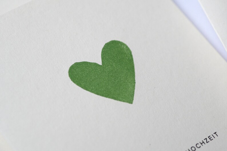 May include: A white card with a green heart in the center. The word "HOCHZEIT" is printed in black at the bottom of the card.