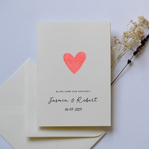 May include: A white card with a pink heart and the text "Alles Liebe zur Hochzeit"  "Jasmin & Robert" "06.07.2024".